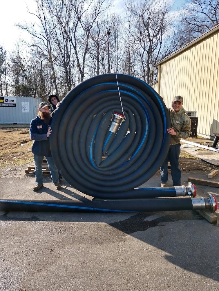 Three B&B Hose team members holding a massive coiled large-bore hose assembly built for Norfolk Naval Shipyard